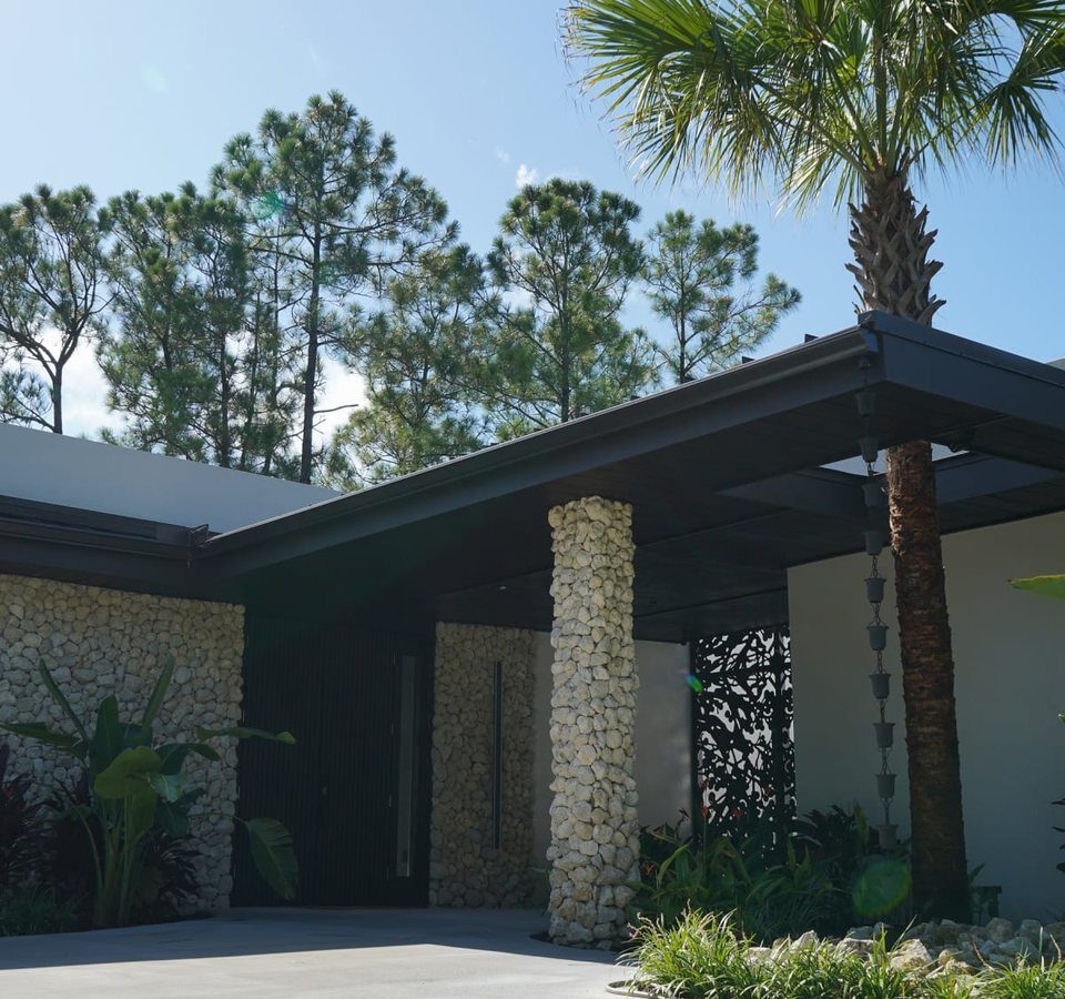 Front of a modern custom home built by Lifestyle Design Homes in Naples, Florida, with a coquina stone wall, a stone-clad column at the entry and a covered walkway beside a palm