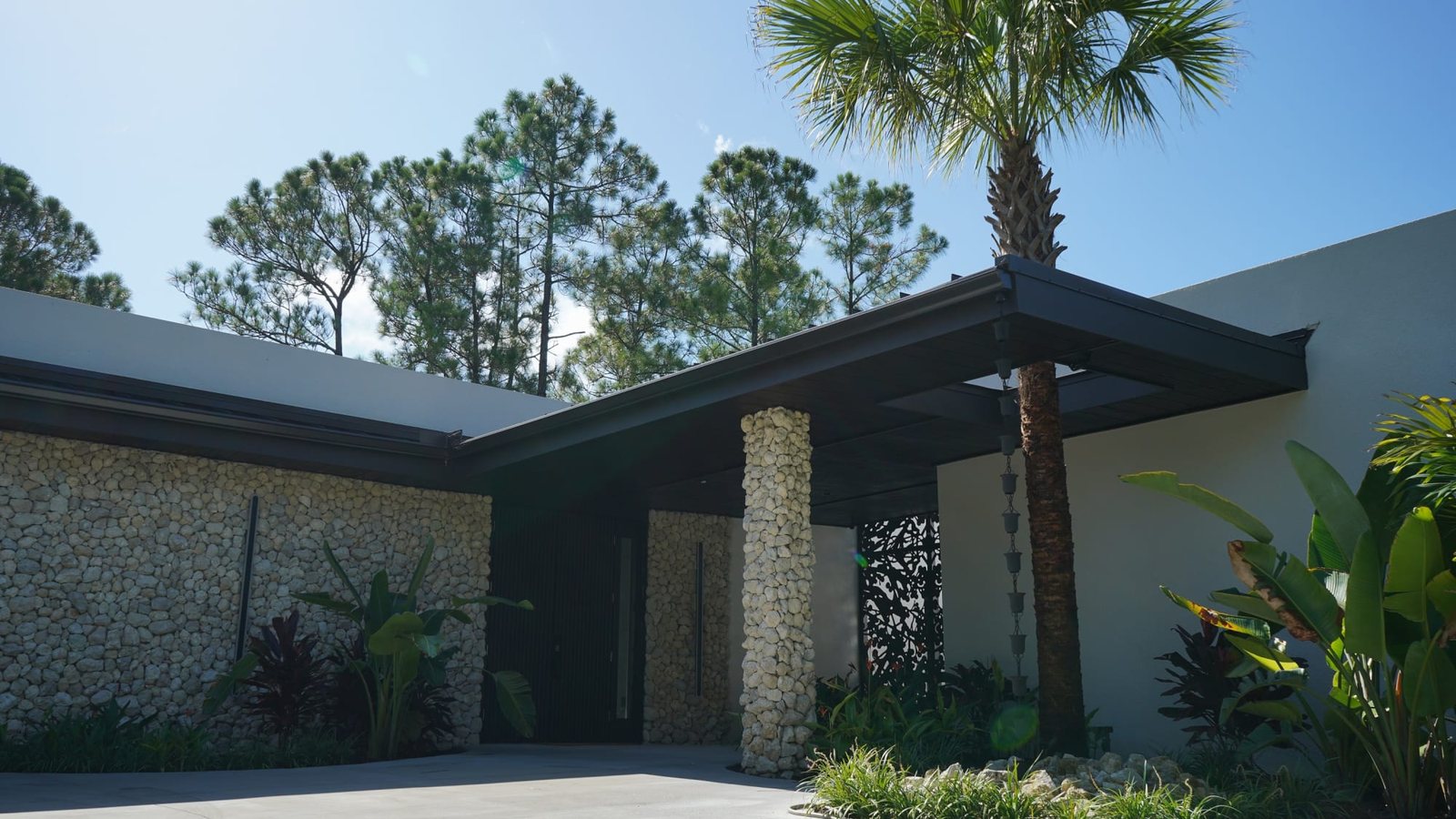 Front of a modern custom home built by Lifestyle Design Homes in Naples, Florida, with a coquina stone wall, a stone-clad column at the entry and a covered walkway beside a palm